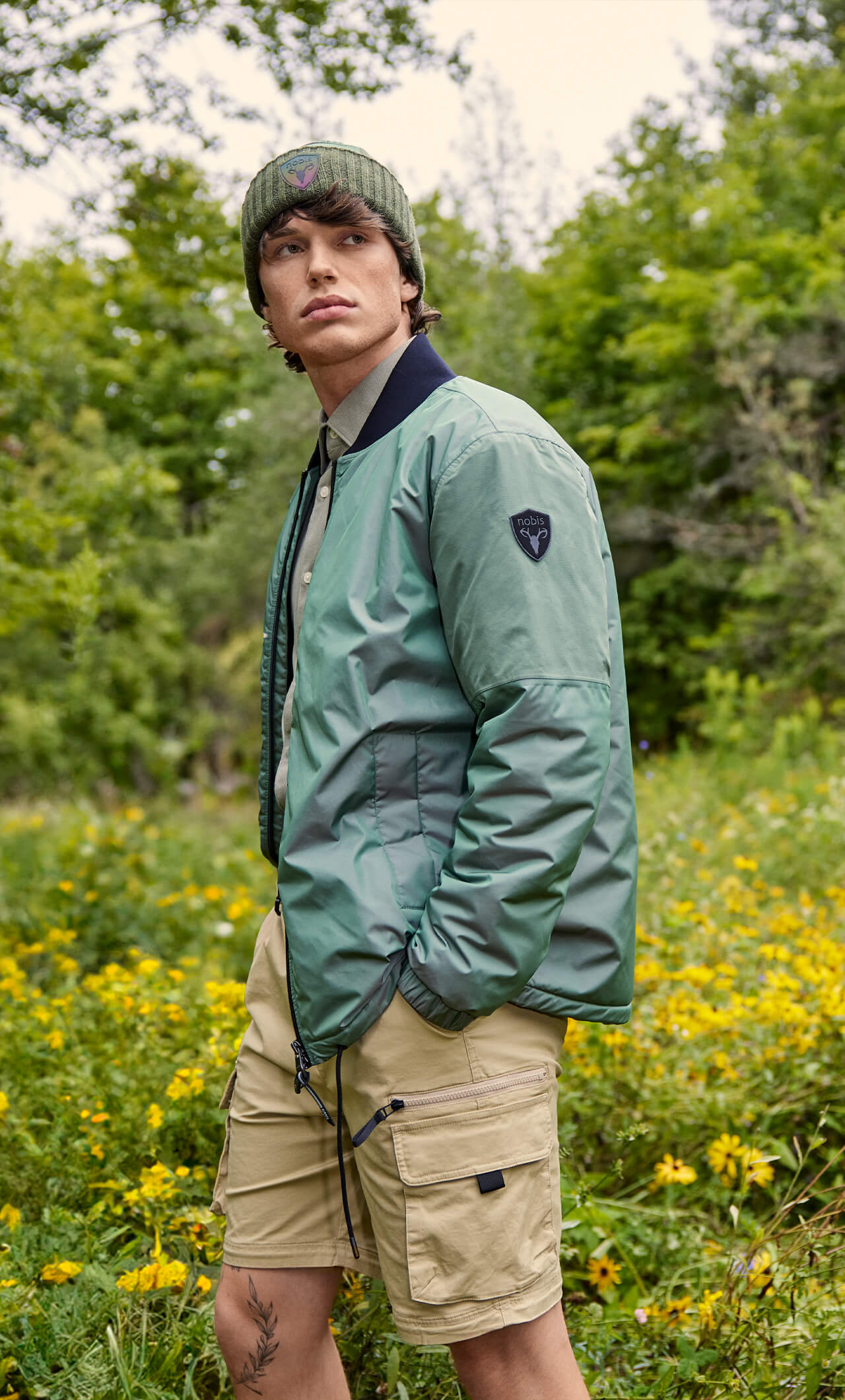 Man standing in a field wearing khaki shorts, a green bomber jacket and a green beanie.