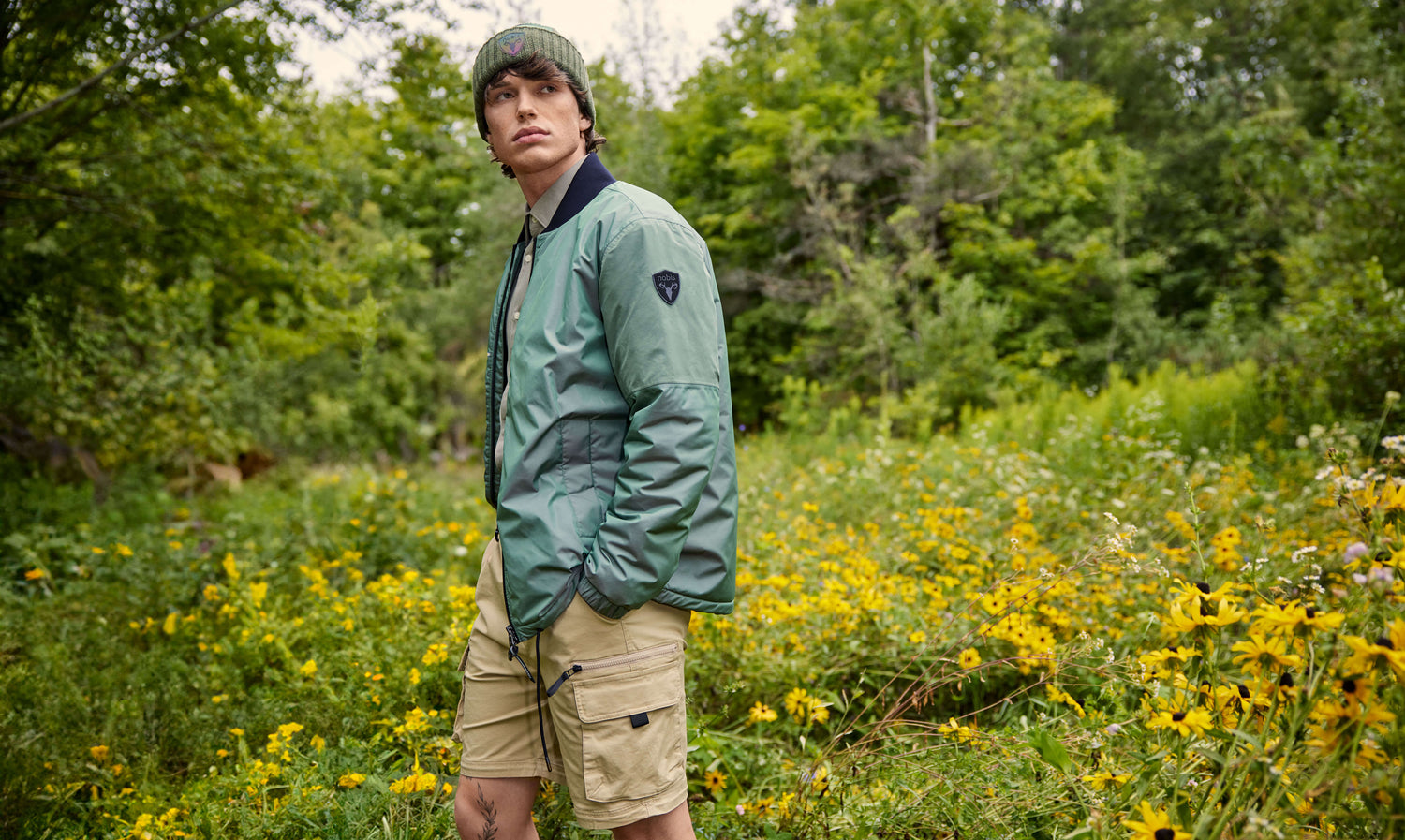 Man standing in a field wearing khaki shorts, a green bomber jacket and a green beanie.