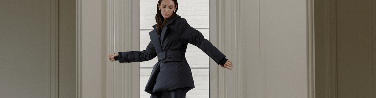 A woman wearing a dark navy blue belted coat stands in a doorway with her arms outstretched, set against a neutral beige interior background.