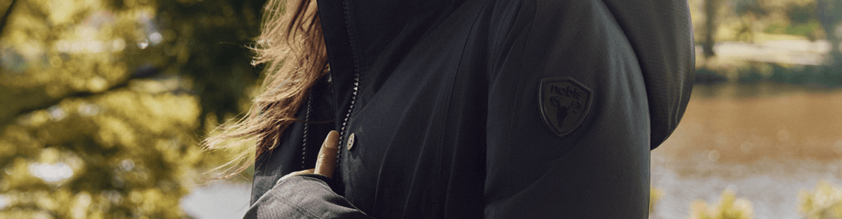 A close-up of a person wearing a black hooded jacket outdoors, with blurred trees and water in the background. A shield-shaped logo patch is visible on the upper sleeve.