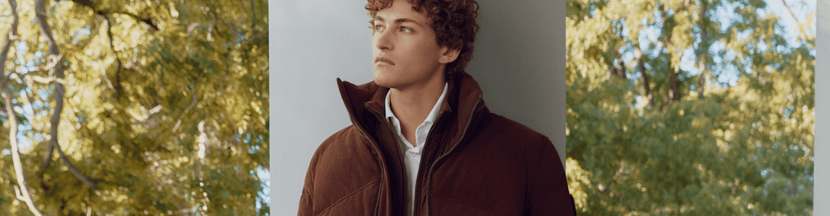 A man wearing a dark brown puffer jacket over a white collared shirt, standing outdoors with blurred green trees on both sides.