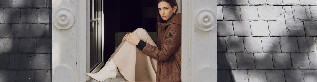 A model wearing a brown quilted jacket and beige trousers sits in a doorway between gray shingled walls.
