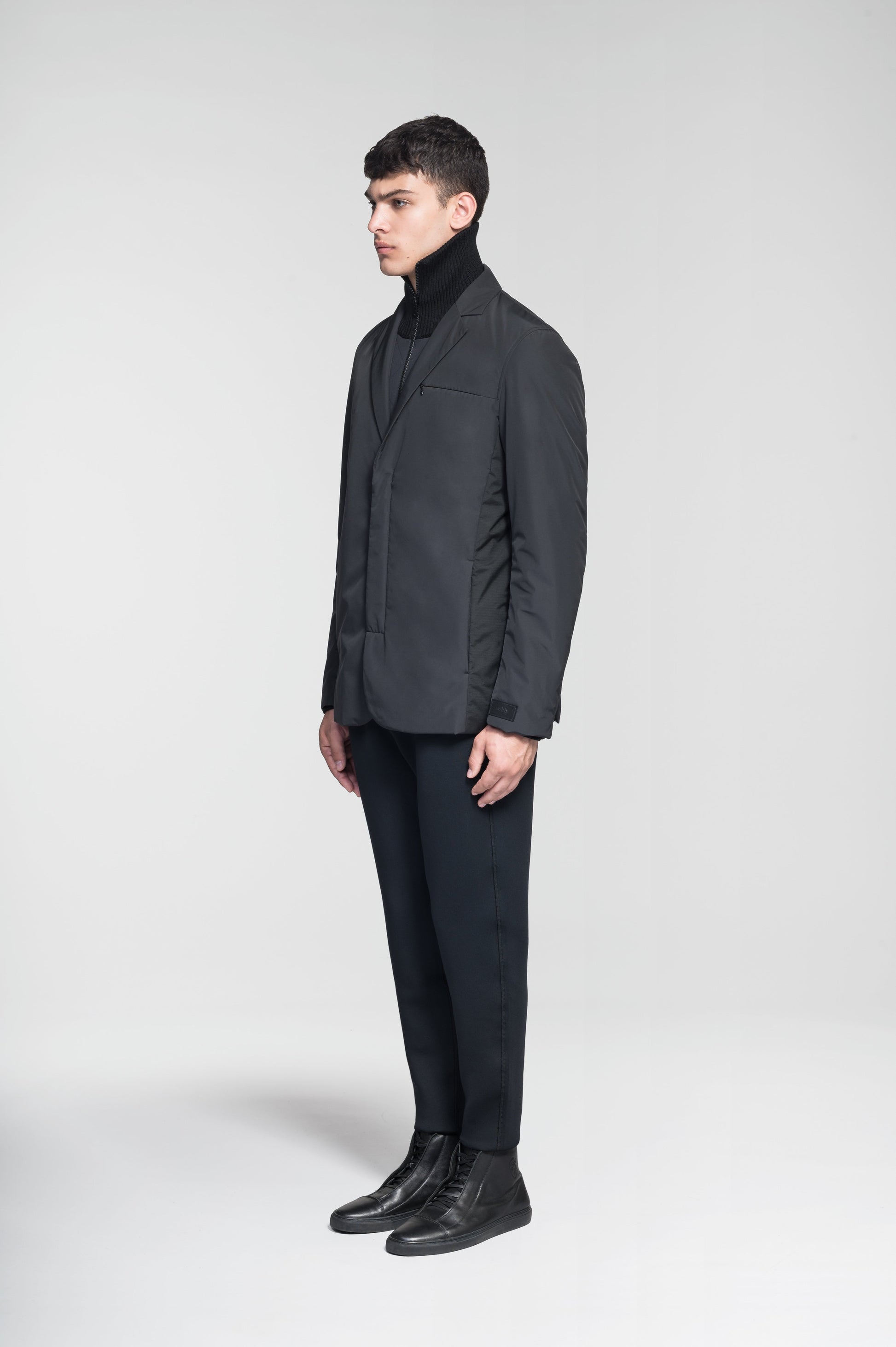 Cody Men's Tailored Travel Blazer in 3-ply micro denier and stretch nylon fabrication with DWR coating, Primaloft Gold Insulation Active+, hidden two-way zipper at centre front with snap closure placket, three invisible exterior zipper pockets, double back pleats, and hidden snap placket at cuffs, in Black