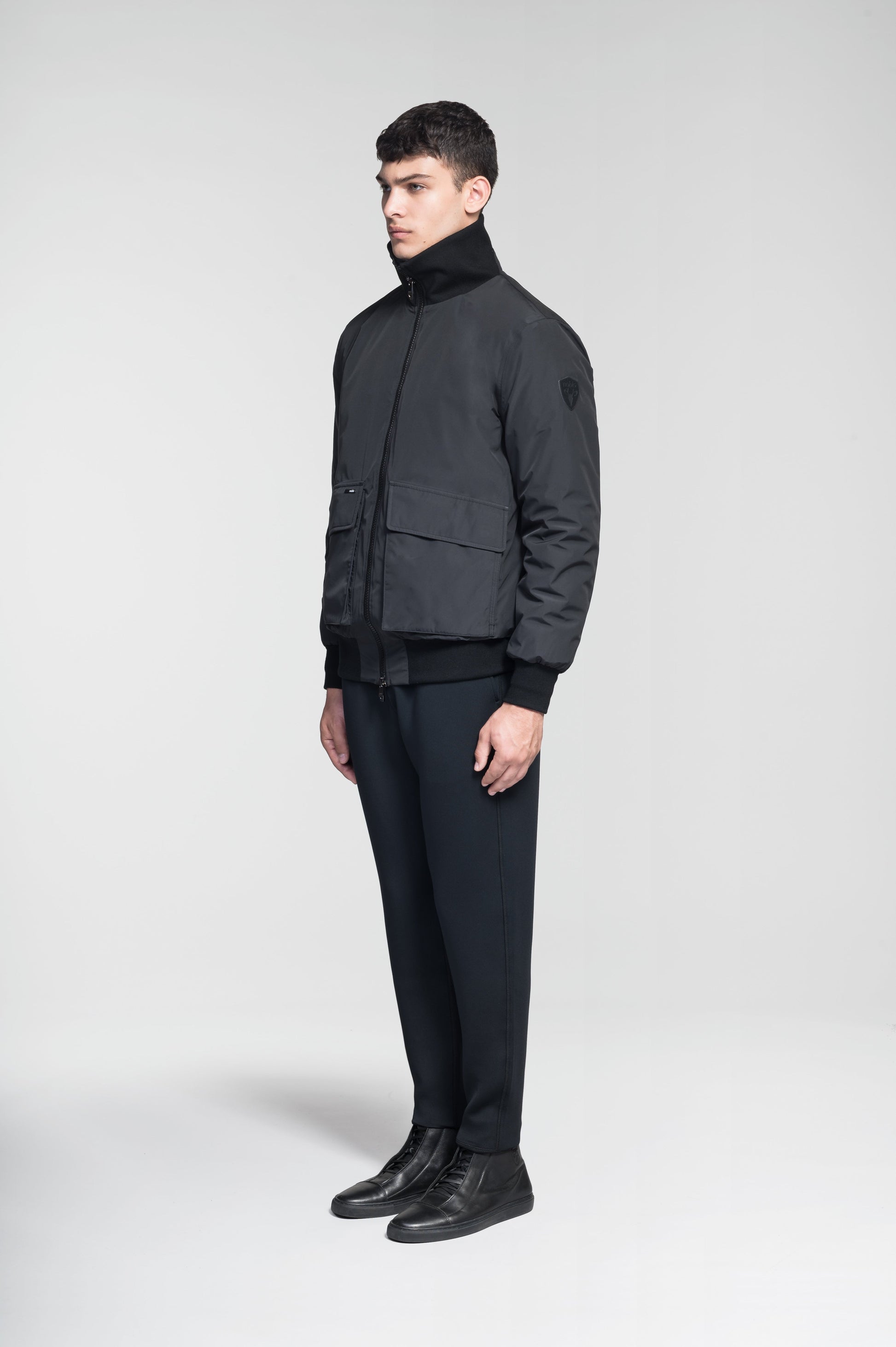 Flint Men's Tailored Rib Collar Jacket in hip length, premium 3-ply Micro Denier fabrication, premium 4-way stretch, water resistant Primaloft Gold Insulation Active +, flap pockets with magnetic closure at waist, side entry pockets at waist, ribbed sleeve cuffs, two-way branded zipper at centre front, box pleat detailing at centre back, large interior zipper pocket, and interior button pocket at left chest, in Black